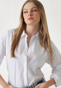 Camicia bianca con bottoni, caratterizzata da piccoli e discreti borchie decorative, maniche lunghe e vestibilità rilassata, abbinata a jeans in denim.
