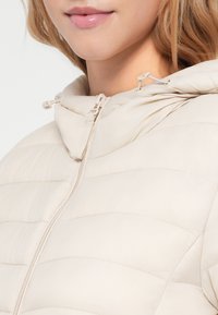 Beige gewatteerde jas met een hoge kraag, voorzien van horizontale stiksels, een voormodelrits en trekkoordverstellingen bij de kap.