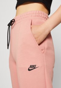 Růžové tepláky s elastickým páskem, černou šňůrkou na stažení, bočními kapsami a černým logem Nike na stehně. Měkká textura.