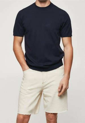 Hombre con una camiseta azul marino lisa de manga corta y pantalones vaqueros hasta la rodilla de color blanco roto, con una mano en el bolsillo, frente a un fondo neutro.