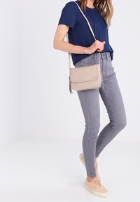 Beżowa listonoszka wykonana z miękkiego materiału, z klapką na zakładkę, detalami przeszyć i regulowanym paskiem. Noszona z szarymi dżinsami i trampkami.