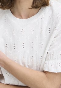 Top in cotone bianco con un motivo testurizzato, dettagli in pizzo e maniche corte a volant. La scollatura è rotonda e rilassata.