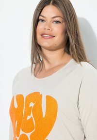 Beige sweatshirt med en stor, texturerad orange grafik som säger "HEY!" med avslappnad passform och rund halsringning.