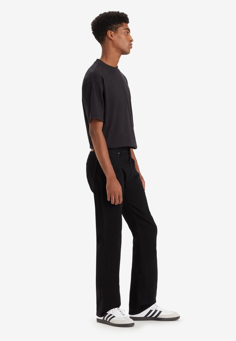 Schwarzes T-Shirt mit kurzen Ärmeln, kombiniert mit schwarzen geraden Jeans und weißen Sneakers mit schwarzen Akzenten. Einfaches Design, glatte Textur.