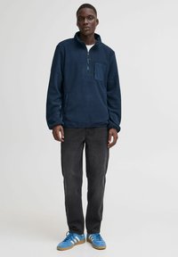 Pullover in pile blu navy con chiusura a mezza zip e tasca frontale. Abbinato a jeans neri e sneakers blu con dettagli bianchi.