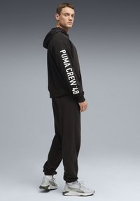 Sweat à capuche noir avec "PUMA CREW '48" en blanc sur la manche, associé à un pantalon de survêtement noir assorti et des baskets grises avec des accents verts.