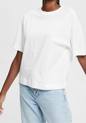 Personne portant un t-shirt blanc uni à manches courtes et ample, et un jean en denim bleu clair, debout avec les bras légèrement étendus.