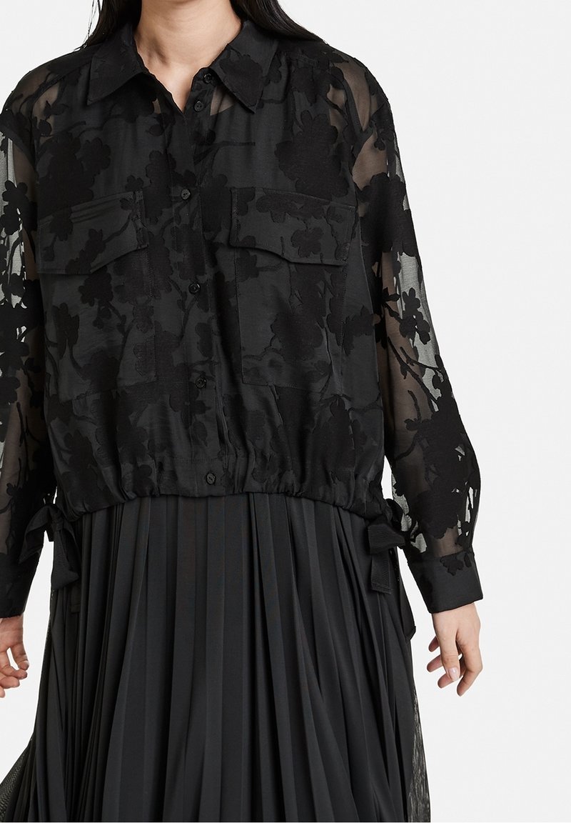 Femme portant un chemisier noir transparent brodé de fleurs avec boutons et deux poches poitrine, ainsi qu'une jupe noire plissée.