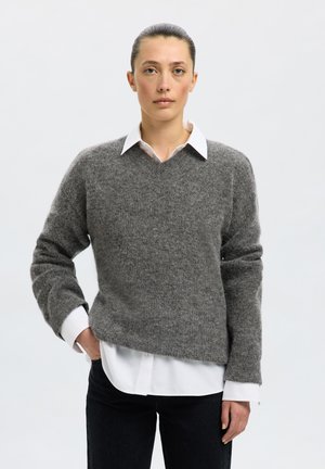 Femme aux cheveux foncés attachés porte un pull gris par-dessus une chemise blanche à col et un pantalon noir, debout avec une main dans la poche.