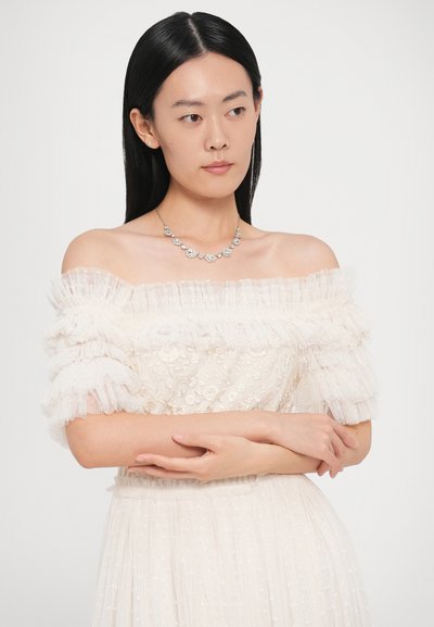 Kremowa sukienka z odkrytymi ramionami, haftem kwiatowym, warstwowymi falbanami na rękawach oraz teksturowaną spódnicą. Do tego srebrny naszyjnik w kształcie kwiatów.