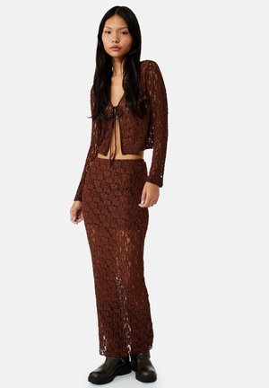 Mujer con cabello largo y oscuro que lleva un conjunto marrón de dos piezas de crochet con mangas largas y falda de talle alto combinada con botas negras.