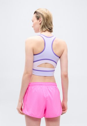 Femme aux cheveux blonds courts portant un débardeur de sport lavande à dos nageur et un short de sport rose vif, debout devant un fond blanc uni.