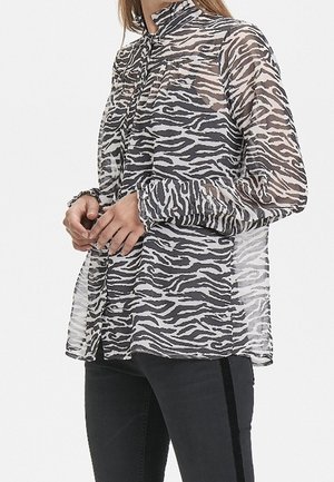 Kobieta w przezroczystej bluzce z długim rękawem o czarno-białym paskowanym wzorze zebry, zestawiona z czarnymi spodniami z bocznym paskiem.