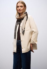 Cremefarvet jakke med knaplukninger, to forlommer og løse ærmer. Sammen med blå jeans og et mønstret brunt og sort tørklæde.