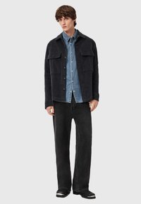 Černá strukturovaná bunda se dvěma předními kapsami, přes světle modrou džínovou košili, spárovaná s volnými černými džíny a černými botami.