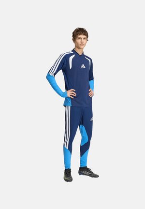 Jeune homme debout, mains sur les hanches, portant une tenue de sport Adidas bleue et bleu clair avec des crampons noirs sur un fond uni.