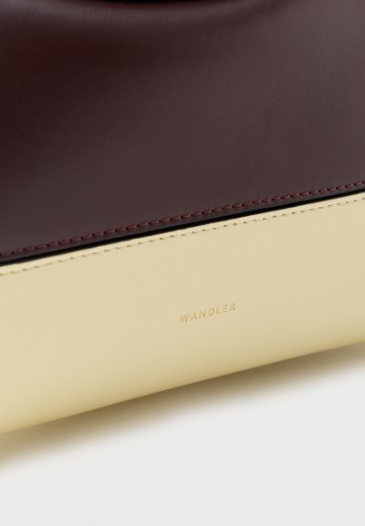 Κοντινή λήψη ενός δίχρωμου δερμάτινου τσάντας με σκούρα βυσσινί επάνω και κρεμ χαμηλότερα πάνελ, με χρυσό λογότυπο "WANDLER" στην κρεμ περιοχή.