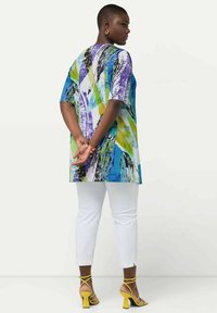 Túnica colorida de manga corta con un patrón geométrico en azul, verde y púrpura; combinada con pantalones blancos y sandalias amarillas de tacón.