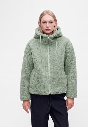 Jeune personne aux cheveux clairs portant une veste polaire zippée à capuche vert clair et un pantalon foncé, debout devant un fond uni.