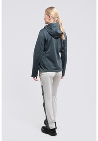 Softshell-Jacke in Dunkelgrün, kombiniert mit hellbeigen Hosen. Die Hosen verfügen über dunkelgrüne Akzente. Beide haben eine glatte, dehnbare Textur.