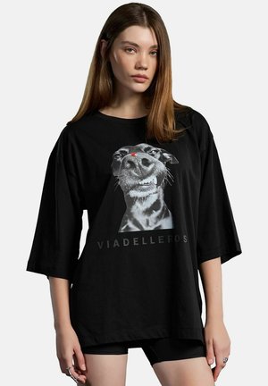 Jeune femme aux cheveux bruns raides portant un T-shirt noir oversize avec un graphique en gros plan du visage d'un chien et le texte "VIADELLEROS".