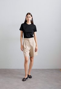 Woman wearing black short-sleeve shirt, beige high-waisted knee-length shorts, black woven flats, and dangling earrings against plain wall.