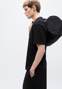 Μαύρο κοντομάνικο μπλουζάκι, συνδυασμένο με φαρδιά μαύρα παντελόνια. Μία κυλινδρική μαύρη τσάντα ακουμπά στον ώμο, τονίζοντας από λεπτομέρεια χρυσού κρίκου.