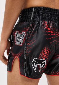 Fekete és piros kígyómintás sportnadrág elasztikus derékrésszel, oldalsó sliccel, valamint fehér kígyófog- és "MV" logókkal a jobb combon.