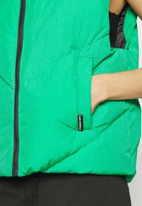 Groene gewatteerde bodywarmer met een rits aan de voorkant, zijzak en een klein zwart label. Het materiaal heeft een gladde textuur met een lichte glans.
