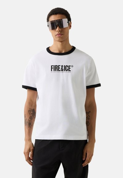 Mand iført hvid T-shirt med sort kant og teksten "FIRE & ICE", sorte bukser, store futuristiske solbriller og synlige tatoveringer på armene.