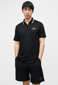 Μαύρο πόλο μπλουζάκι με φερμουάρ, χρυσές λεπτομέρειες και λογότυπο. Συνδυάζεται με μαύρο σορτς με υφή και πλαϊνές τσέπες.