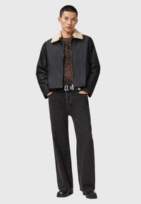 Chaqueta bomber negra con un collar de borreguito crema, cierre de cremallera, bolsillos laterales, usada sobre una blusa con estampado y jeans anchos negros.