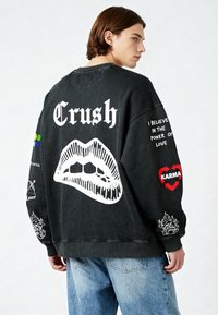Felpa nera oversize con la parola "Crush" in bianco, una grafica di labbra e vari patch colorati sulle maniche. Texture invecchiata.