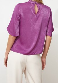Blusa corta viola a maniche corte con una finitura morbida e testurizzata e un dettaglio a chiave sulla schiena. Abbinata a pantaloni a gamba larga di colore crema.