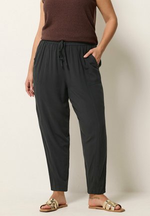 Kobieta w czarnych spodniach z sznurkiem, brązowym topie bez rękawów i złotych sandałach wsuwanych, stoi z jedną ręką w kieszeni.