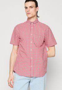 Chemise à manches courtes à carreaux rouges et blancs avec col boutonné et poche poitrine, présentant une coupe décontractée et un tissu léger.