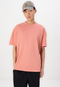 Jasnoróżowa bawełniana koszulka z luźnym krojem, okrągłym dekoltem i krótkimi rękawami, w połączeniu z wyblakłą szaro-czarną czapką z logo.