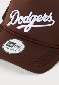 Căciulă de baseball maro, ajustabilă, cu text alb brodat "Dodgers" și un autocolant circular al brandului New Era pe margine.