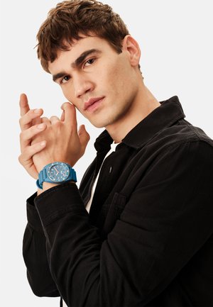 Blåt ur med rund urskive og gummirem, båret på en mandlig håndled. Manden har en sort jakke over en hvid skjorte på og poserer tænksomt.