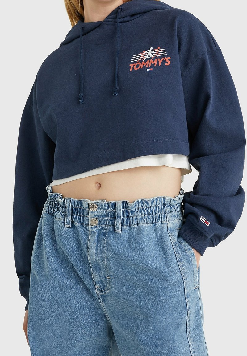 Sudadera recortada de color navy con logotipo, cordones y mangas largas, combinada con unos jeans de tiro alto en un tono azul claro, con cintura elástica y botones.