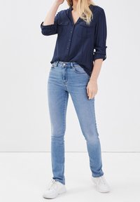 Chemise marine boutonnée avec deux poches avant, associée à un jean skinny bleu clair et des baskets blanches, avec les manches retroussées.