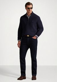 Tengerészkék cipzáras pulóver, alatta csíkos galléros ing, sötét farmer, barna cipő és napszemüveg. Egyszerű kialakítás bordázott mandzsettával és szegéllyel.