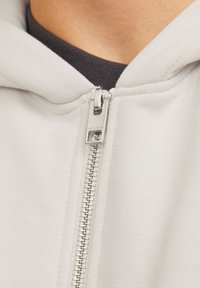 Sweatshirt à capuche gris clair avec une fermeture éclair en argent, texture côtelée près du col et une couche intérieure gris foncé visible en dessous.
