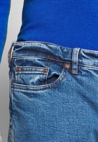 Jean en denim bleu clair avec une texture délavée, présentant des coutures orange et une petite poche avant sur le côté droit, porté à la taille.