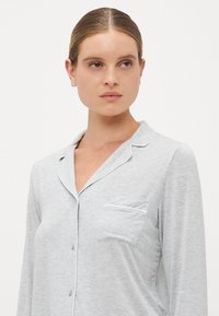 Kvinna med slickat hår som bär en ljusgrå knappad pyjamasöverkropp med krage och bröstficka, som ser åt sidan.
