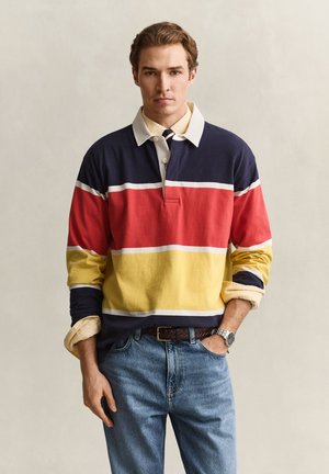 Jeune homme portant un maillot de rugby rayé multicolore, une chemise claire, un jean et une montre, debout devant un fond uni.