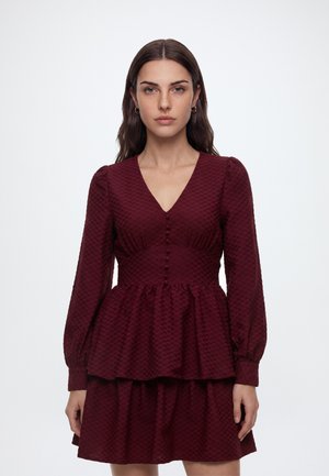 Femme aux longs cheveux foncés porte une robe bordeaux texturée avec manches longues, col en V, boutons couverts et taille à basque.