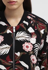 Nærbillede af en person iført en sort hættetrøje med lynlås, dekoreret med blomsterdesign i rød, hvid og grøn samt ribkant.
