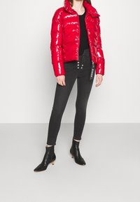 Červená lesklá péřová bunda s vysokým límcem, zapínáním na zip a štítkem značky. V kombinaci s černými slim kalhotami a černými kotníkovými botami.