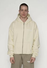 Lysegul hættejakke med lynlås i tekstureret stof, med en frontlomme og "MULTIPLY"-logo i hvid. Kombineret med grå bukser.